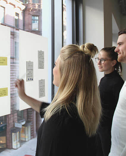 Team aus drei Personen sammelt Ideen in Form von Klebezetteln an einer Glaswand.